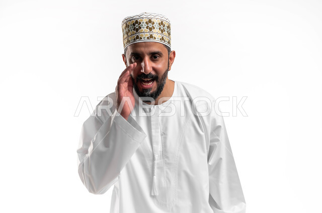 الكلام والنداء والتحدث بصوت مرتفع، بورتريه مقرب لشاب عربي خليجي عماني مبتسم يرتدي الدشداشة و الكمه ينظر الى الكاميرا يضع يده على فمه بإيماءات المناداة لشخص ما، دردشة ومحادثات وتواصل اجتماعي، خلفية بيضاء