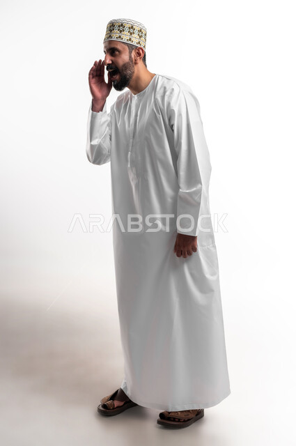 النداء والكلام والتحدث بصوت مرتفع، بورتريه لشاب عربي خليجي عماني مبتسم يرتدي الدشداشة و الكمه يضع يده على فمه بإيماءات المناداة لشخص ما، دردشة ومحادثات وتواصل اجتماعي، صورة الجسم بالطول الكامل, خلفية بيضاء