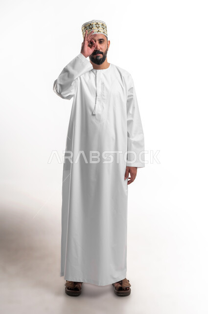 مفهوم التخطيط و الرؤية المستقبلية، بورتريه لشاب عربي خليجي عماني مبتسم يرتدي الدشداشة و الكمه يضع يده على عينيه بشكل عدسة بإيماءات البحث والاستكشاف، صورة الجسم بالطول الكامل, خلفية بيضاء