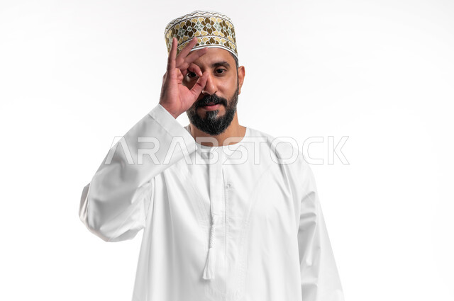 مفهوم التخطيط و الرؤية المستقبلية، بورتريه مقرب لشاب عربي خليجي عماني مبتسم يرتدي الدشداشة و الكمه يضع يده على عينيه بشكل عدسة بإيماءات البحث والاستكشاف، خلفية بيضاء