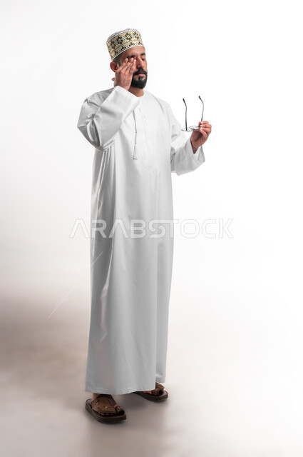 ارتداء النظارات الخاصة بضعف النظر، بورتريه لشاب عربي خليجي عماني مبتسم يرتدي الدشداشة و الكمه يمسك النظاره الطبيه يضع يده على وجهه بإيماءات التعب والإرهاق، مفهوم تعديل وتحسين الرؤية، صورة الجسم بالطول الكامل, خلفية بيضاء