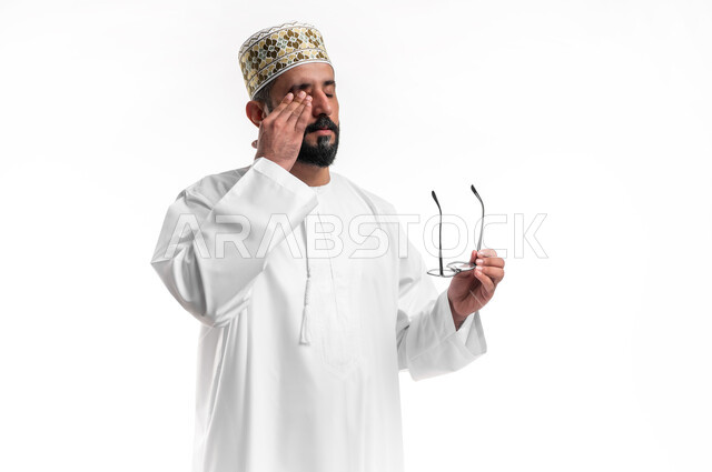 ارتداء النظارات الخاصة بضعف النظر، بورتريه مقرب لشاب عربي خليجي عماني مبتسم يرتدي الدشداشة و الكمه يمسك النظاره الطبيه يضع يده على وجهه بإيماءات التعب والإرهاق، مفهوم تعديل وتحسين الرؤية، خلفية بيضاء