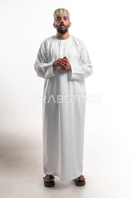 الاستغراب من أمر معين، الشعور بالصدمة والذهول، بورتريه لشاب عربي خليجي عماني مبتسم يرتدي الدشداشة و الكمه ينظر الى الكاميرا بإيماءات أيدي متماسكة، مفهوم التعجب والاندهاش من شيء ما، صورة الجسم بالطول الكامل, خلفية بيضاء