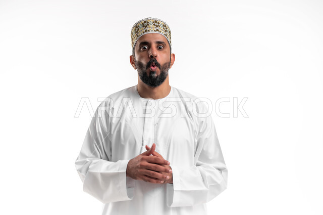 الاستغراب من أمر معين، الشعور بالصدمة والذهول، بورتريه مقرب لشاب عربي خليجي عماني مبتسم يرتدي الدشداشة و الكمه ينظر الى الكاميرا بإيماءات أيدي متماسكة، مفهوم التعجب والاندهاش من شيء ما، خلفية بيضاء
