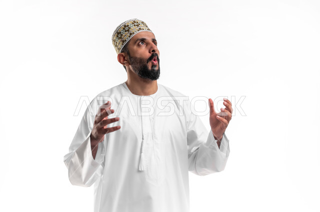 الشعور بالصدمة والذهول، الاستغراب من أمر معين، بورتريه مقرب لشاب عربي خليجي عماني مبتسم يرتدي الدشداشة و الكمه ينظر الى الاعلى يرفع يديه بإيماءات التعجب والاندهاش من شيء ما، خلفية بيضاء