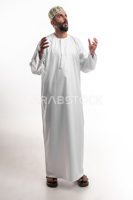 الشعور بالصدمة والذهول، الاستغراب من أمر معين، بورتريه مقرب لشاب عربي خليجي عماني مبتسم يرتدي الدشداشة و الكمه ينظر الى الاعلى يرفع يديه بإيماءات التعجب والاندهاش من شيء ما، صورة الجسم بالطول الكامل, خلفية بيضاء