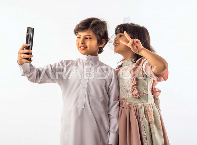 خلفية بيضاء لصبي وفتاه صغيران سعوديان مبتسمان ، يمسك الصبي الجوال بيده , يقومان بإلتقاط صور سيلفي تذكاريه بإستخدام الهاتف المحمول بإيماءات وجه ويدين مختلفة , بورتريه لطفل وطفلة سعوديان خليجيان سعيدان يرتديان لباس العيد ,الاحتفال بعيد الفطر السعيد