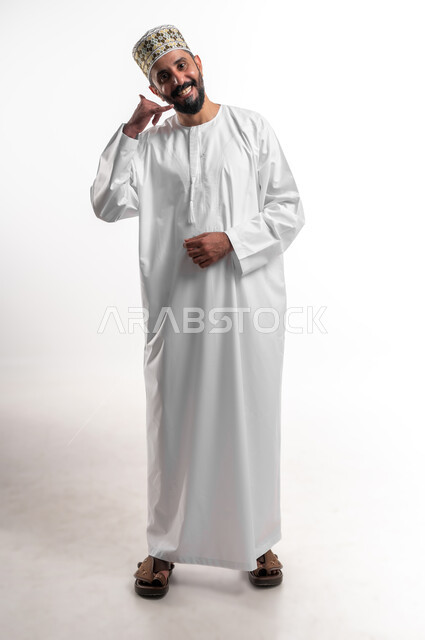 إنتظار مكالمة وإستخدام الميكروفون، بورتريه لشاب عربي خليجي عماني مبتسم يرتدي الدشداشة و الكمه يرفع يده بإيماءات الإتصال، إجراء مكالمة هاتفية، تعابير الفرح والسرور، صورة الجسم بالطول الكامل, خلفية بيضاء
