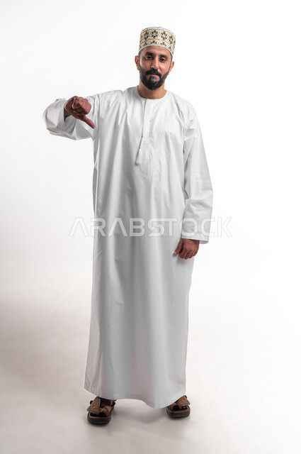 مفهوم الرفض وعدم القبول، بورتريه لشاب عربي خليجي عماني يرتدي الدشداشة و الكمه يشير بإصبع الابهام للاسفل بإيماءات عدم الاعجاب والتقييم السلبي، الشعور بالانزعاج من شيء ما، صورة الجسم بالطول الكامل, خلفية بيضاء