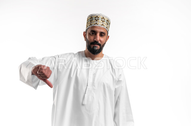 The concept of rejection and non-acceptance, a close-up portrait of a young Arab Gulf Omani man wearing a dishdasha and a cuff, pointing with his thumb down with gestures of disliking and negative evaluation, feeling annoyed by something, white background