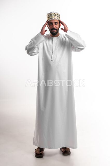 مفهوم الحاجة الى الهدوء، بورتريه لشاب عربي خليجي عماني يرتدي الدشداشة و الكمه والنظارة الطبية يضع يديه على رأسه بتعابير التفكير، حل المشكلات واتخاذ القرارات, إيماءات التمعن والتركيز والتخطيط للمستقبل، صورة الجسم بالطول الكامل, خلفية بيضاء