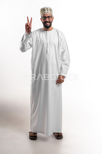 النجاح وتحقيق الأهداف، مفهوم السعادة بالوصول إلى الغايات، بورتريه لشاب عربي خليجي عماني يرتدي الدشداشة و الكمه والنظارة الطبية يرفع يده باشارة النصر والقوة، إيماءات السرور بالفوز والانتصار، صورة الجسم بالطول الكامل, خلفية بيضاء