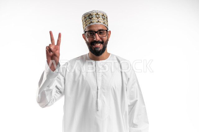 النجاح وتحقيق الأهداف، مفهوم السعادة بالوصول إلى الغايات، بورتريه مقرب لشاب عربي خليجي عماني يرتدي الدشداشة و الكمه والنظارة الطبية يرفع يده باشارة النصر والقوة، إيماءات السرور بالفوز والانتصار، خلفية بيضاء
