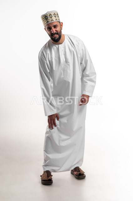 الأوجاع والآلام العصبية والعضلية، طلب المساعده والحاجه للعلاج، التهاب العضلات والمفاصل، بورتريه لشاب عربي خليجي عماني يرتدي الدشداشة و الكمه يضع يده على ركبته بايماءات الألم والتعب والأرهاق، حالة اسعافية طارئة،  صورة الجسم الطول الكامل, خلفية بيضاء