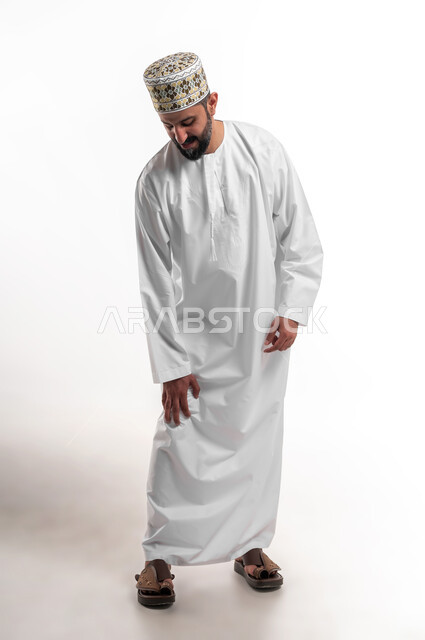الأوجاع والآلام العصبية والعضلية، طلب المساعده والحاجه للعلاج، التهاب العضلات والمفاصل، بورتريه لشاب عربي خليجي عماني يرتدي الدشداشة و الكمه يضع يده على ركبته بايماءات الألم والتعب والأرهاق، حالة اسعافية طارئة،  صورة الجسم الطول الكامل, خلفية بيضاء
