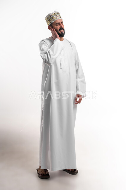 Inflammation of the middle ear, the need to go to the doctor and develop an appropriate treatment plan, portrait of a young Arab Gulf Omani man wearing a dishdasha and a cuff, placing his hand on his ear with gestures of extreme pain, an emergency emergency, full-length body portrait, white background