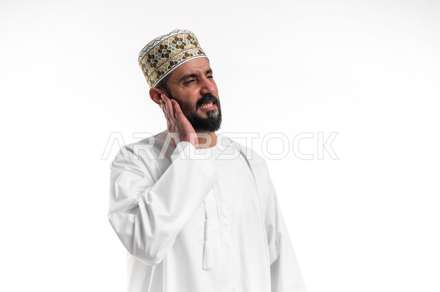التهاب الأذن الوسطى، الحاجة للذهاب للطبيب و وضع خطة علاجية مناسبة، بورتريه مقرب لشاب عربي خليجي عماني يرتدي الدشداشة و الكمه يضع يده على اذنه بايماءات التألم الشديد، حالة اسعافية طارئة،  خلفية بيضاء
