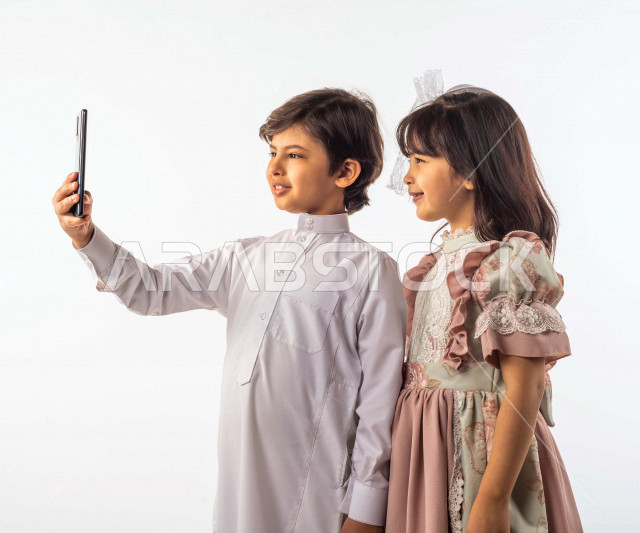 White background of two smiling Saudi boy and girl, boy holding mobile phone in hand, taking memorial selfies using mobile phone with different face and hand gestures, portrait of a happy Gulf Saudi boy and girl wearing Eid clothes, celebrating Eid al-Fit