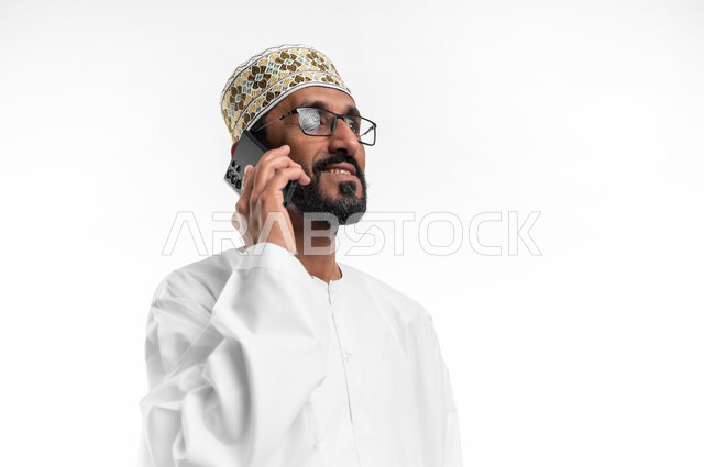 الدردشة مع الأهل والأصدقاء، تسهيل التواصل مع الآخرين باستخدام الهاتف المحمول، تطورات تقنية وتكنولوجية، بورتريه لرجل عماني شايب يرتدي الدشداشة والكمه يقوم بالمحادثة مع الأقارب عبر الموبايل، خلفية بيضاء