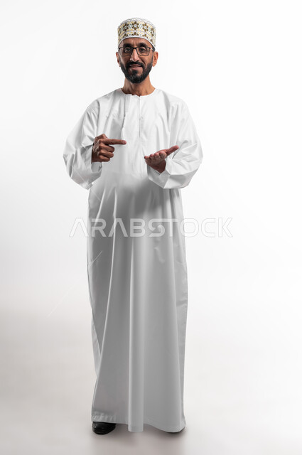 النظر إلى الكاميرا بايماءات السعادة و السرور، رفع شيء ما في كف والاشارة إليه في الكف الآخر، بورتريه لرجل عربي عماني شايب يرتدي الدشداشة والكمه يشير بإصبعه إلى عنصر ما، تعابير فرح و رضا، خلفية بيضاء
