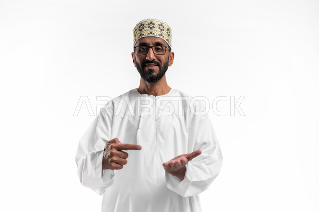 النظر إلى الكاميرا بايماءات السعادة و السرور، رفع شيء ما في كف والاشارة إليه في الكف الآخر، بورتريه لرجل عربي عماني شايب يرتدي الدشداشة والكمه يشير بإصبعه إلى عنصر ما، تعابير فرح و رضا، خلفية بيضاء