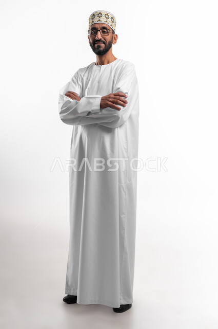 الوقوف باستقامة مع تقاطع الايدي، بورتريه لرجل عربي عماني شايب يرتدي الدشداشة والكمه يقف بشكل متكتف ينظر إلى الكاميرا بتعابير الابتسامة، وضع اليدين على الذراعين وشد الظهر، خلفية بيضاء