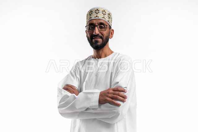 الوقوف باستقامة مع تقاطع الايدي، بورتريه لرجل عربي عماني شايب يرتدي الدشداشة والكمه يقف بشكل متكتف ينظر إلى الكاميرا بتعابير الابتسامة، وضع اليدين على الذراعين وشد الظهر، خلفية بيضاء