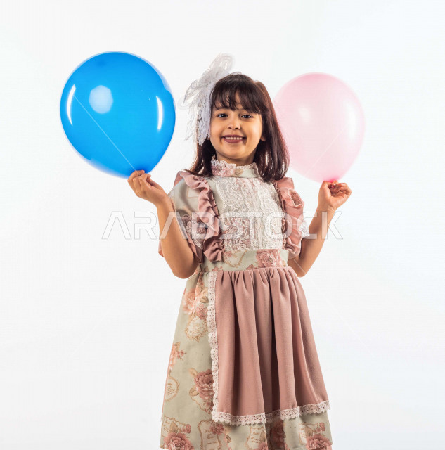 خلفية بيضاء لفتاه صغيرة سعودية خليجية مبتسمة وجميلة ، تنظر الى الكاميرا وتحمل بيدها الأولى بالون أزرق وبيدها الأخرى زهري بإيماءات وجه ويدين مختلفة , بورتريه لطفلة سعودية خليجية سعيدة ترتدي لباس العيد الانيق, الاحتفال بعيد الفطر السعيد ،عيد الفطر المبارك