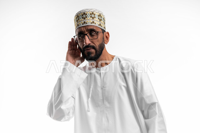 وضع اليد خلف الأذن ومحاولة رصد الصوت القادم، بورتريه لرجل عربي عماني شايب يرتدي الدشداشة والكمه يحاول سماع الأصوات من حوله، حدوث ضعف في السمع مع التقدم في السن، علامات الكبر في العمر، خلفية بيضاء