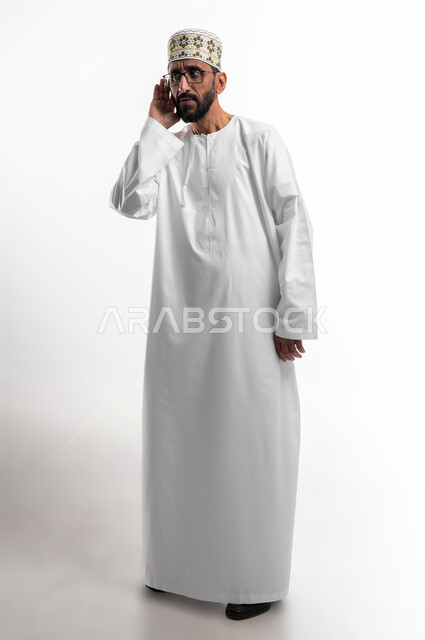 وضع اليد خلف الأذن ومحاولة رصد الصوت القادم، بورتريه لرجل عربي عماني شايب يرتدي الدشداشة والكمه يحاول سماع الأصوات من حوله، حدوث ضعف في السمع مع التقدم في السن، علامات الكبر في العمر، خلفية بيضاء