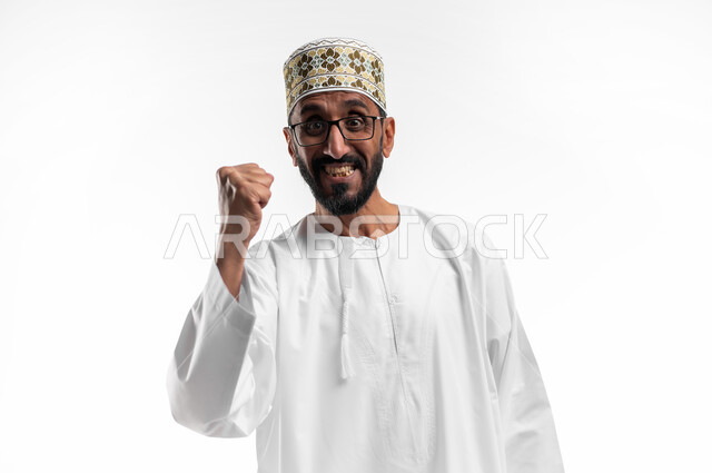 ايماءات وجه تدل على الحماس الزائد، بورتريه لرجل عربي عماني شايب يرتدي الدشداشة والكمه يرفع يده للأعلى للتشجيع، تعابير فرح وسرور، الافتخار والشعور بالسعادة لإنجاز شيء ما، خلفية بيضاء