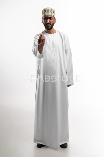 A hand movement means lowering the volume and calming down, facial and hand gestures indicating an attempt to calm a heated discussion, a portrait of an old Omani Arab man wearing a dishdasha and a cuff, showing signs of annoyance and discomfort, the way to ask politely and respectfully, a white background.