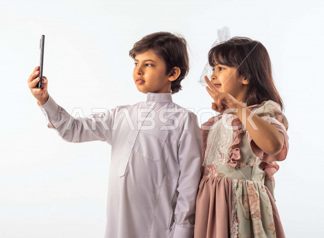 خلفية بيضاء لصبي وفتاه صغيران سعوديان مبتسمان ، يمسك الصبي الجوال بيده , يقومان بإلتقاط صور سيلفي تذكاريه بإستخدام الهاتف المحمول بإيماءات وجه ويدين مختلفة , بورتريه لطفل وطفلة سعوديان خليجيان سعيدان يرتديان لباس العيد ,الاحتفال بعيد الفطر السعيد