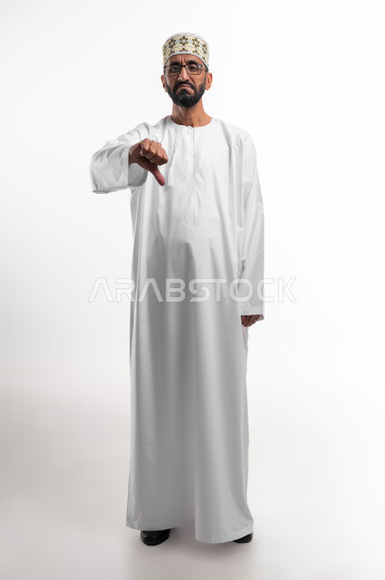 حركة يد وايماءات وجه تدل على عدم الإعجاب والموافقة، بورتريه لرجل عربي عماني شايب يرتدي الدشداشة والكمه تظهر عليه علامات عدم القبول، الإشارة بإصبع الإبهام للاسفل دليل الرفض، خلفية بيضاء
