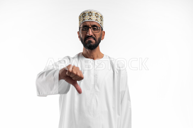 حركة يد وايماءات وجه تدل على عدم الإعجاب والموافقة، بورتريه لرجل عربي عماني شايب يرتدي الدشداشة والكمه تظهر عليه علامات عدم القبول، الإشارة بإصبع الإبهام للاسفل دليل الرفض، خلفية بيضاء
