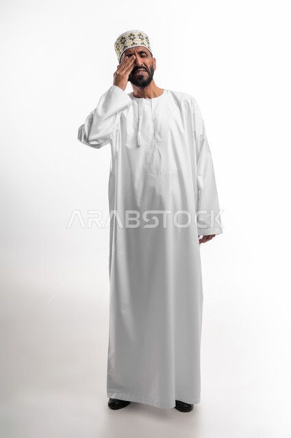 ايماءات وجه ويدين تدل على دخول شي مزعج ومؤلم في العين، بورتريه لرجل عربي عماني شايب يرتدي الدشداشة والكمه يضع يده على عينه تظهر عليه علامات الألم، عدم القدرة على فتح العينين، خلفية بيضاء