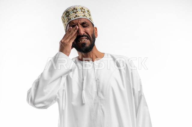 ايماءات وجه ويدين تدل على دخول شي مزعج ومؤلم في العين، بورتريه لرجل عربي عماني شايب يرتدي الدشداشة والكمه يضع يده على عينه تظهر عليه علامات الألم، عدم القدرة على فتح العينين، خلفية بيضاء