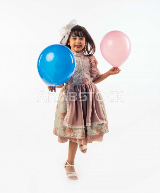 خلفية بيضاء لفتاه صغيرة سعودية خليجية مبتسمة وجميلة ، تنظر الى الكاميرا وتحمل بيدها الأولى بالون أزرق وبيدها الأخرى زهري بإيماءات وجه ويدين مختلفة , بورتريه لطفلة سعودية خليجية سعيدة ترتدي لباس العيد الانيق, الاحتفال بعيد الفطر السعيد ،عيد الفطر المبارك