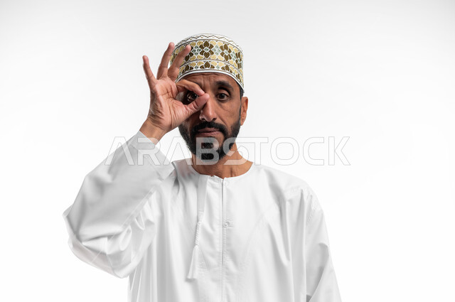 مفهوم التخطيط و الرؤية المستقبلية، بورتريه لرجل عربي عماني شايب يرتدي الدشداشة والكمه يضع يده على عينيه بشكل عدسة بإيماءات البحث والاستكشاف، خلفية بيضاء