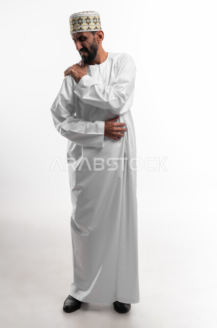 Feeling severe pain that is difficult to bear in the shoulder, difficulty moving and folding the arm, a portrait of an old Omani man wearing a dishdasha and a cuff suffering from pain in his joints, facial gestures indicating pain, a white background.