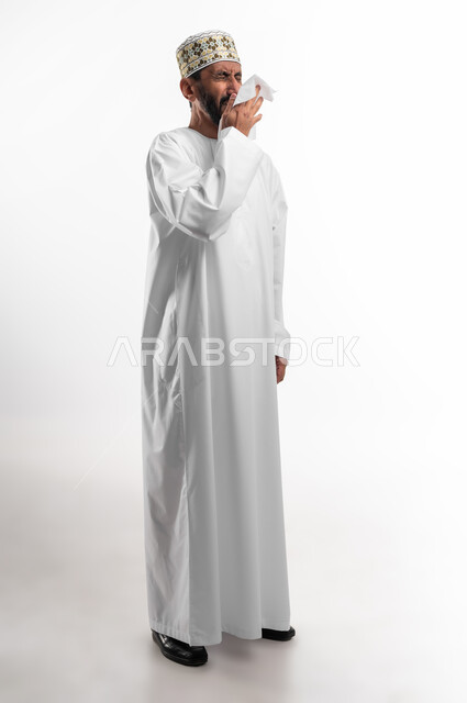 (Covid-19) Corona symptoms: Getting a cold, portrait of an Omani Gulf Arab man wearing a sleeve and a dishdasha, using paper tissues while sneezing, full length body, white background
