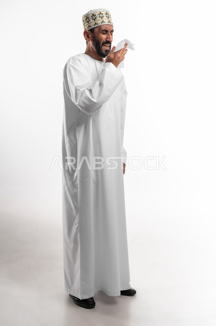 الإصابة بفايروس كورونا كوفيد19،الانفلونزا والتهاب الرئة، الشعور بالمرض والكحة, الإحساس بالكسل والخمول، بورتريه لرجل عربي خليجي عماني يرتدي الدشداشة و الكمه يمسك بيده منديل، استعمال المناديل الورقية أثناء العطس، الجسم بالطول الكامل، خلفية بيضاء