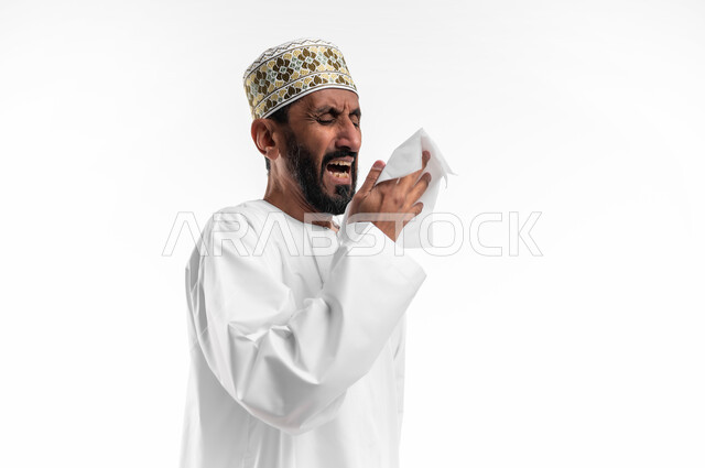 اعراض كورونا (كوفيد-19) الإصابة بنزلة برد, بورتريه مقرب لرجل عربي خليجي عماني يرتدي الكمه والدشداشة يقوم باستخدام المناديل الورقيه أثناء العطاس, خلفية بيضاء