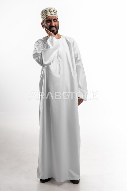 الاحساس بالوجع نتيجة تسوس الاضراس و التهاب اللثه، الحاجة لزيارة الطبيب، طلب العلاج والمساعدة،  بورتريه لرجل عربي خليجي عماني يرتدي الدشداشة والكمه يضع يده على خده ويغمض عينيه بايماءات الشعور بألم الأسنان، الجسم بالطول الكامل، خلفية بيضاء
