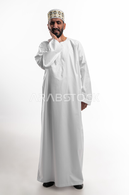 الحاجة لزيارة الطبيب، طلب العلاج والمساعدة، تسوس الاضراس و التهاب اللثه،  بورتريه لرجل عربي خليجي عماني يرتدي الدشداشة والكمه يضع يده على خده ويغمض عينيه بايماءات الشعور بألم الأسنان، الجسم بالطول الكامل، خلفية بيضاء