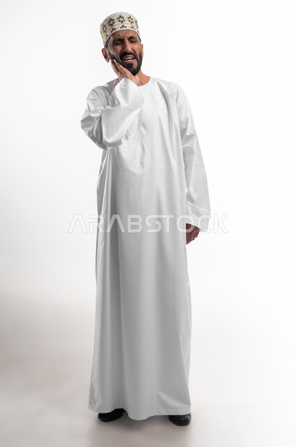 طلب العلاج والمساعدة، تسوس الاضراس و التهاب اللثه،  بورتريه لرجل عربي خليجي عماني يرتدي الدشداشة والكمه يضع يده على خده ويغمض عينيه بايماءات الشعور بألم الأسنان، الحاجة لزيارة الطبيب، الجسم بالطول الكامل، خلفية بيضاء