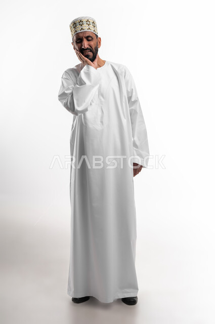 التهاب اللثه وتسوس الاضراس، طلب العلاج والمساعدة، بورتريه لرجل عربي خليجي عماني كبير بالسن يرتدي الدشداشة والكمه يضع يده على خده يغمض عينيه بايماءات الشعور بألم الأسنان، الحاجة لزيارة الطبيب، الجسم بالطول الكامل، خلفية بيضاء