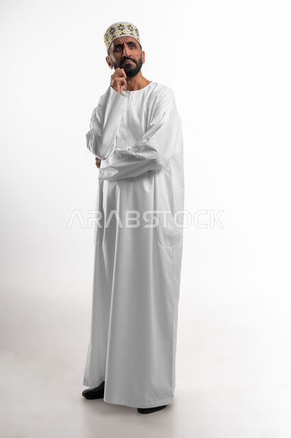 التأمل و التخطيط للمستقبل, بورتريه  لرجل عربي خليجي عماني كبير بالسن يرتدي الدشداشة والكمه يضع يده على ذقنه بايماءات التفكير و التركيز، ايجاد حل لمشكلة ما, تعابير شرود الذهن و التمعن, الجسم بالطول الكامل, خلفية بيضاء