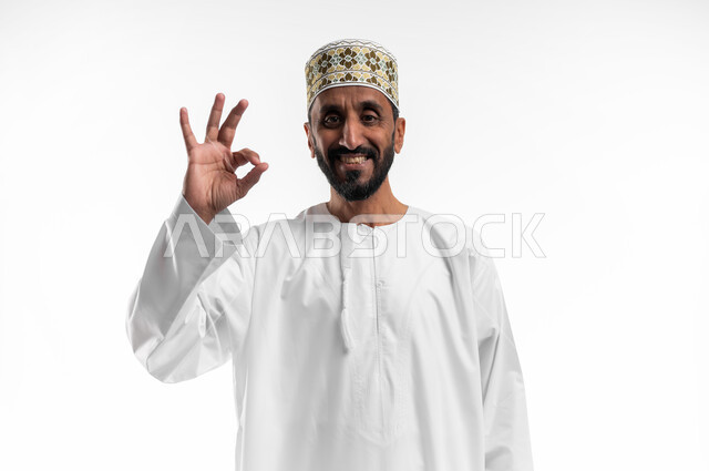 التعبير عن الجودة والإتقان, بورتريه مقرب  لرجل كبير السن عربي خليجي عماني يرتدي الدشداشة والكمه  يرفع يده للأعلى  بإيماءات   الرضا والقبول والأعجاب, النظر الى الكاميرا بتعابير السرور, خلفية بيضاء.