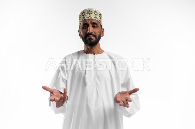 التردد والشعور بالحيرة بأمر معين, بورتريه مقرب لرجل كبير السن  عربي خليجي عماني يرتدي الدشداشة والكمه  برفع كفات يديه للأعلى بإيماءات  عدم المعرفة, الاستغراب وعدم التأكد, خلفية بيضاء.